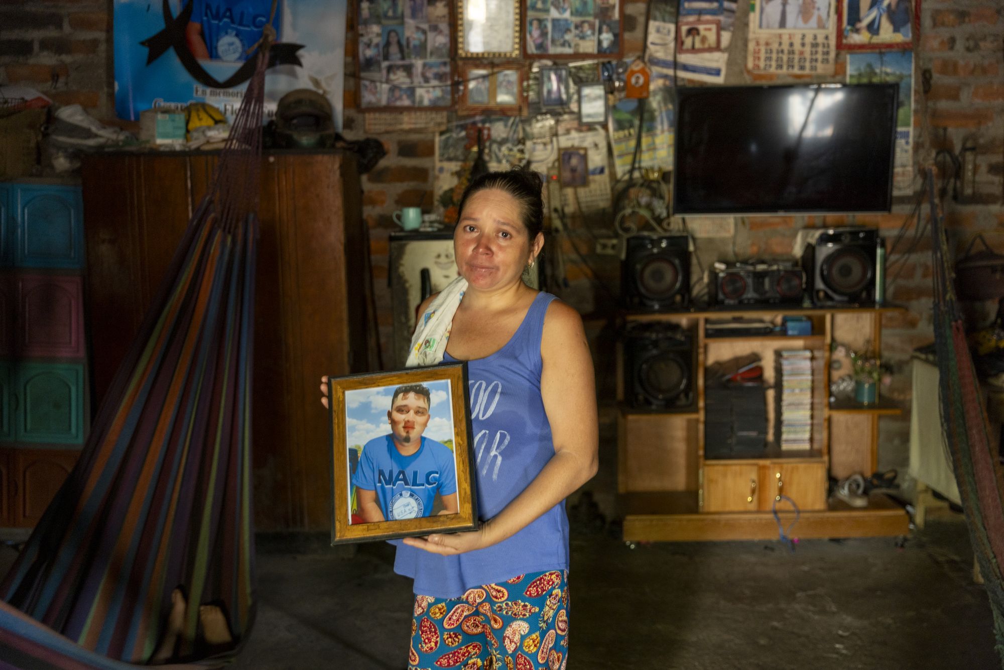 César Omar Flores Fuentes is pictured in a frame held by his mother, Blanca Rosa. Behind her, a memorial banner and family photographs hang on the wall. She is still wrapping her head around the loss of her son.